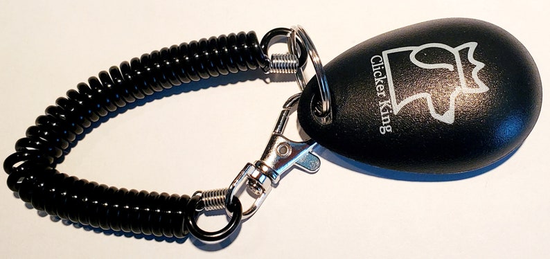 May include: Black clicker with a coiled black cord and a silver clasp. The clicker has the words "Clicker King" and a crown logo. This training tool is designed for pet training.