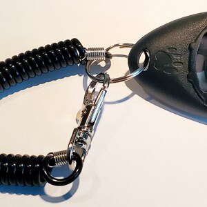 May include: Black pet training clicker with a coiled black cord and a metal clasp. The clicker has a paw print design and a button. The cord is attached to a metal ring.