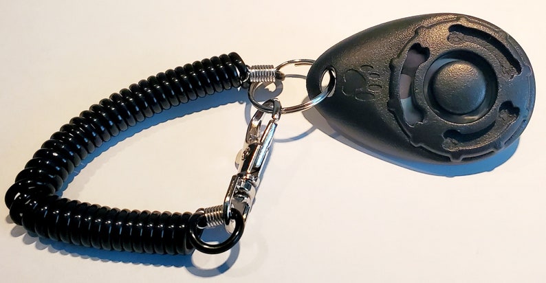May include: Black pet training clicker with a coiled black cord and a metal clasp. The clicker has a paw print design and a button. The cord is attached to a metal ring.
