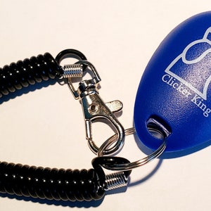 May include: A blue Clicker King dog training clicker with a silver metal clasp and a black coiled wrist strap. The clicker has a white graphic of a dog's head with a crown.
