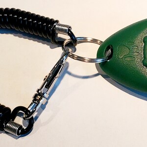 May include: A green pet training clicker with a black button, connected to a black coiled cord and a metal clip. The clicker has a paw print design and is attached to a metal ring.