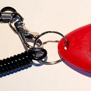 May include: A red dog training clicker with a black button and a black coiled wrist strap. The clicker is attached to a silver metal clasp and a key ring. The clicker is designed for positive reinforcement training.