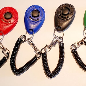 May include: Four dog training clickers in red, blue, black, and green. Each clicker has a black button, a metal keyring, and a black coiled wrist strap with a metal clasp. The clickers are designed for pet training.