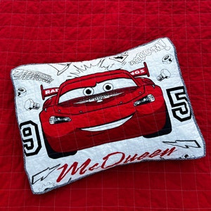 Op de afbeelding: Een wit kussen met een rode cartoon raceauto erop. De auto heeft een glimlachend gezicht en de woorden "McQueen" en "95" staan op het kussen gedrukt. Het kussen heeft een grijze rand.