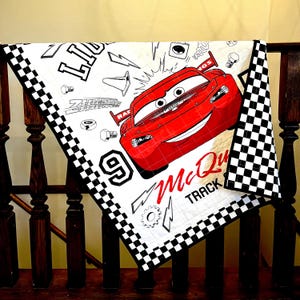 Könnte beinhalten: Eine weiße Steppdecke mit einem großen roten Cartoon-Rennwagen und dem Text "McQueen Track". Die Steppdecke hat einen schwarz-weiß karierten Rand und verschiedene Cartoon-Grafiken.