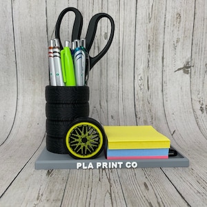 May include: A desk organizer with a tire-themed pen holder, a wheel-shaped accent, and a tray holding sticky notes. The pen holder is made of stacked black tires. The wheel has a yellow rim. The text "PLA PRINT CO" is visible.