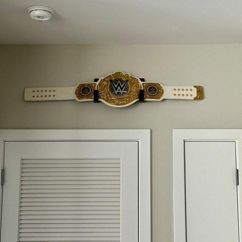 May include: A gold and white wrestling championship belt with the WWE logo, mounted on a wall. The belt has a large central plate and two side plates, all with gold detailing. The white strap has gold studs.