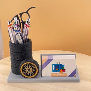 May include: A desk organizer set featuring a pen holder made of stacked black tires, a decorative wheel, and a business card holder. The pen holder contains several pens. The business card holder has a graphic of a 3D printer and the text "PLA PRINT CO".
