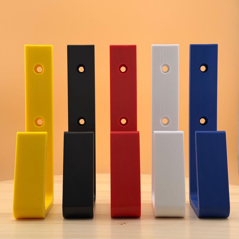May include: A collection of six wall hooks in various colors: yellow, black, red, white, and blue. Each hook has a rectangular back with a circular hole for mounting and a curved, open-ended lower section. The hooks are arranged on a wooden surface.