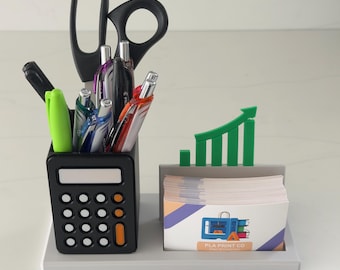 May include: A black calculator-shaped desk organizer with pens, scissors, and a green arrow-shaped accent. The organizer holds business cards with the text "PLA PRINT CO". The desk organizer is on a gray tray.