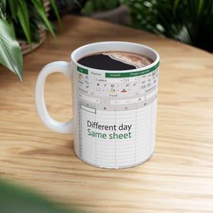 May include: White ceramic coffee mug with a handle, featuring an Excel spreadsheet design. The mug has the text "Different day, Same sheet" printed on it. The mug is filled with dark coffee.