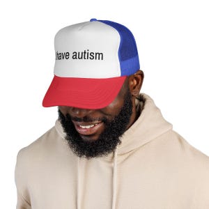 Może przedstawiać: Czapka typu trucker z białym panelem przednim, czerwonym daszkiem i niebieskim tyłem z siatki. Tekst "I have autism" jest wydrukowany na czarno na przednim panelu. Czapkę nosi osoba.