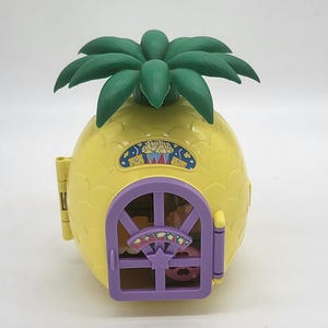 May include: A yellow pineapple-shaped playhouse with a green palm tree roof and a purple door. The door has a window with a star and a small pink and white toy inside.