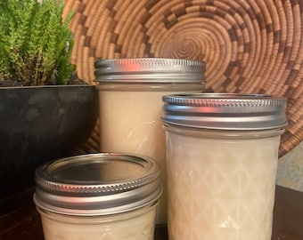 May include: Three glass jars with silver lids filled with white candle wax. The jars are of different sizes and are sitting on a wooden surface.