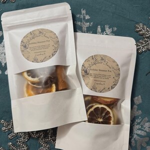 May include: Two white pouches with clear windows, each containing dried orange slices and spices. The pouches have circular labels that read "Holiday Simmer Pot" and "Wildflower Farm". The background is a teal fabric with silver snowflake accents.