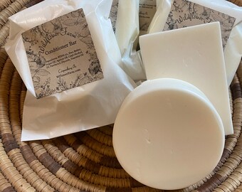 May include: A set of three white conditioner bars wrapped in white paper with a brown label that reads "Wildflower Farm Conditioner Bar" and lists ingredients. The bars are in a woven basket.