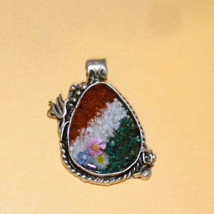 May include: A teardrop-shaped silver pendant featuring a textured metal border with a decorative side accent. The center inlay displays a layered landscape design with brown, white, and green granular textures, accented by two small artificial flowers.