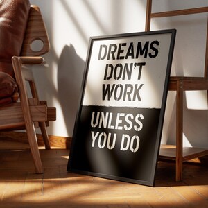 May include: A framed poster with the text "DREAMS DON'T WORK UNLESS YOU DO" in a stencil font. The top half of the poster is white, and the bottom half is black. The poster is leaning against a wall near a wooden chair and a wooden shelf.