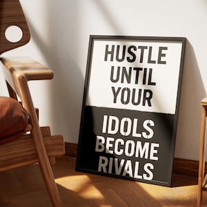 May include: Black framed poster with the text "HUSTLE UNTIL YOUR IDOLS BECOME RIVALS" in white. The poster is leaning against a white wall, next to a wooden chair with a brown leather seat.