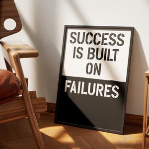 May include: A framed poster with the text "SUCCESS IS BUILT ON FAILURES" in a stencil font. The top half of the poster is white, and the bottom half is black. The poster is leaning against a white wall, next to a wooden chair.