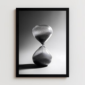 May include: Framed black and white print featuring an hourglass. The hourglass contains dark sand, with the sand flowing from the top to the bottom bulb. The frame is black, and the background is a grey gradient.