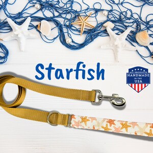 May include: A gold dog leash with a starfish print and a silver clasp. The leash is on a white wooden surface with a blue fishing net and seashells. The text "Starfish" is written in blue letters above the leash. A red, white, and blue label with the text "All Products Handmade in the USA" is in the upper right corner of the image.