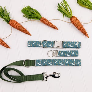 May include: Two dog collars and a matching leash with a teal blue background and a white bunny rabbit and carrot pattern. The collars and leash are on a white surface with a string of carrots with green tops.