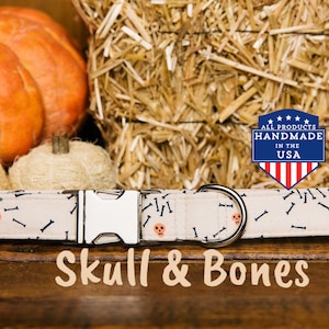 May include: A white dog collar with a skull and bone pattern. The collar has a silver buckle and a silver D-ring. The collar is handmade in the USA. "Skull & Bones" is written in orange text below the collar.