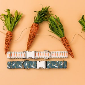 May include: Two dog collars with different patterns. The top collar has a white background with orange carrots printed on it. The bottom collar has a blue background with white rabbits printed on it. Both collars have silver buckles and are displayed against a light orange background with a string of orange carrot decorations.