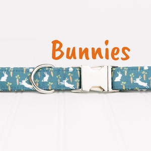 May include: A blue dog collar with a white bunny and carrot pattern. The collar has a silver buckle and a silver D-ring. The text "Bunnies" is written in orange above the collar.