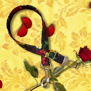 May include: A floral patterned dog leash with a seatbelt buckle closure and a silver-colored metal clasp. The leash is black with red, yellow, and white flowers. A red rose and petals are in the image.