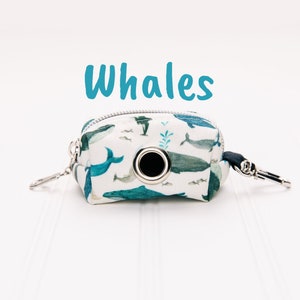 May include: A white poop bag dispenser with a blue whale print and the word "Whales" above it. The dispenser has a silver zipper and a silver clip.