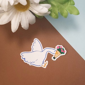 Pode incluir: Um adesivo de desenho animado de ganso branco com contornos azuis segurando um buqu&ecirc; de flores rosa em um cone de papel marrom.