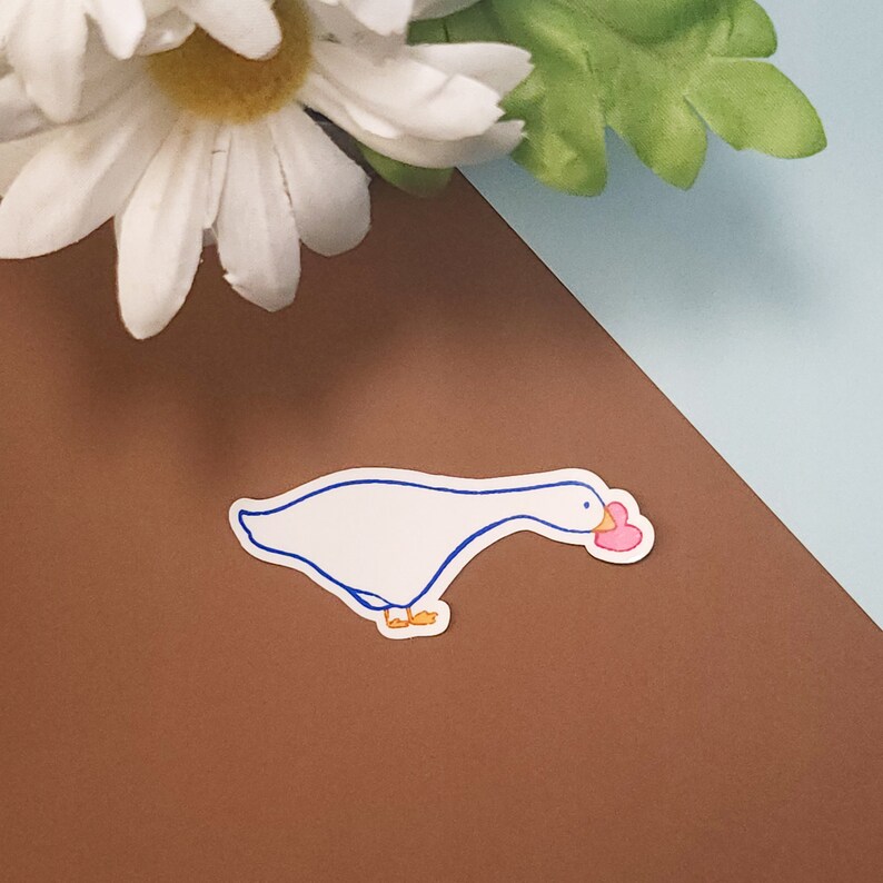 Pode incluir: Um adesivo de ganso branco com contornos azuis, segurando um cora&ccedil;&atilde;o rosa no bico. O adesivo est&aacute; em um fundo marrom.