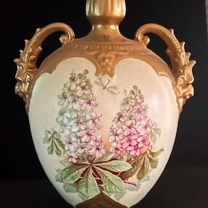 May include: A ceramic vase with a cream-colored body featuring painted pink and white horse chestnut flower clusters and green leaves. The vase has a gold-painted neck, base, and two ornate, scrolled handles on the sides with a metallic finish.