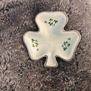 May include: A white, shamrock-shaped dish with a gold trim and green clover accents. The dish is made of ceramic and is a decorative item. The dish is a symbol of St. Patrick's Day.