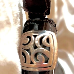 May include: Close-up of a black patent leather handbag with a silver, ornate, square-shaped clasp.