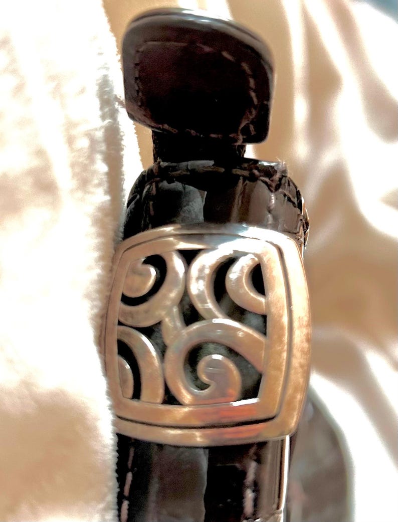 May include: Close-up of a black patent leather handbag with a silver, ornate, square-shaped clasp.