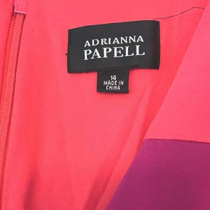 May include: Close-up of a vibrant dress with a color-blocked design. The dress features a coral pink bodice and a deep purple panel. The label reads "ADRIANNA PAPELL" and "14 MADE IN CHINA". A zipper runs down the front.