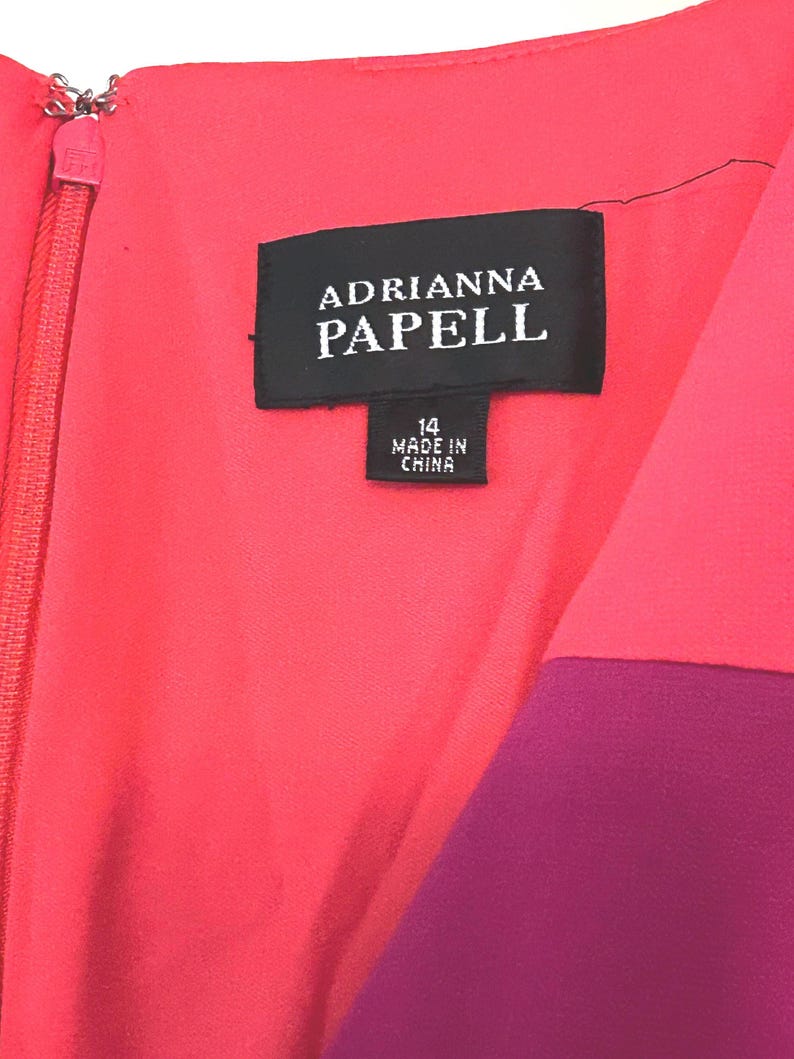 May include: Close-up of a vibrant dress with a color-blocked design. The dress features a coral pink bodice and a deep purple panel. The label reads "ADRIANNA PAPELL" and "14 MADE IN CHINA". A zipper runs down the front.