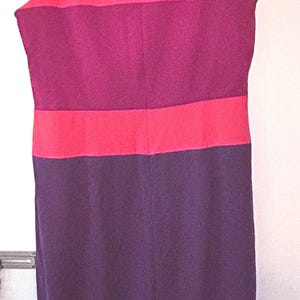 May include: Sleeveless color block dress in shades of pink, purple, and plum. The dress is hanging on a wooden hanger. The top section is pink, followed by a purple band, a pink band, and a plum-colored skirt.