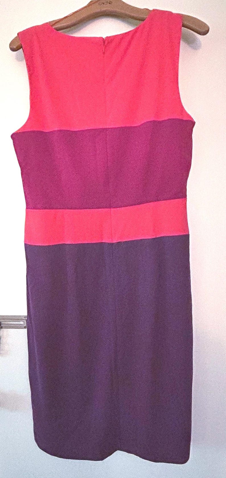 May include: Sleeveless color block dress in shades of pink, purple, and plum. The dress is hanging on a wooden hanger. The top section is pink, followed by a purple band, a pink band, and a plum-colored skirt.