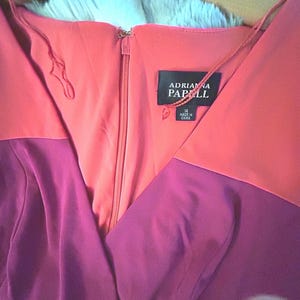 May include: A two-tone dress with a V-neckline, featuring a coral bodice and a deep purple skirt. The dress has a zipper closure and a tag that reads "Adrianna Papell". The dress is hanging on a wooden hanger.