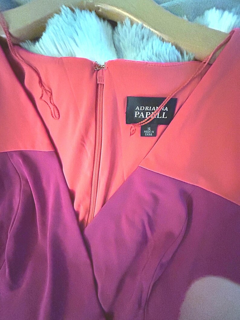 May include: A two-tone dress with a V-neckline, featuring a coral bodice and a deep purple skirt. The dress has a zipper closure and a tag that reads "Adrianna Papell". The dress is hanging on a wooden hanger.