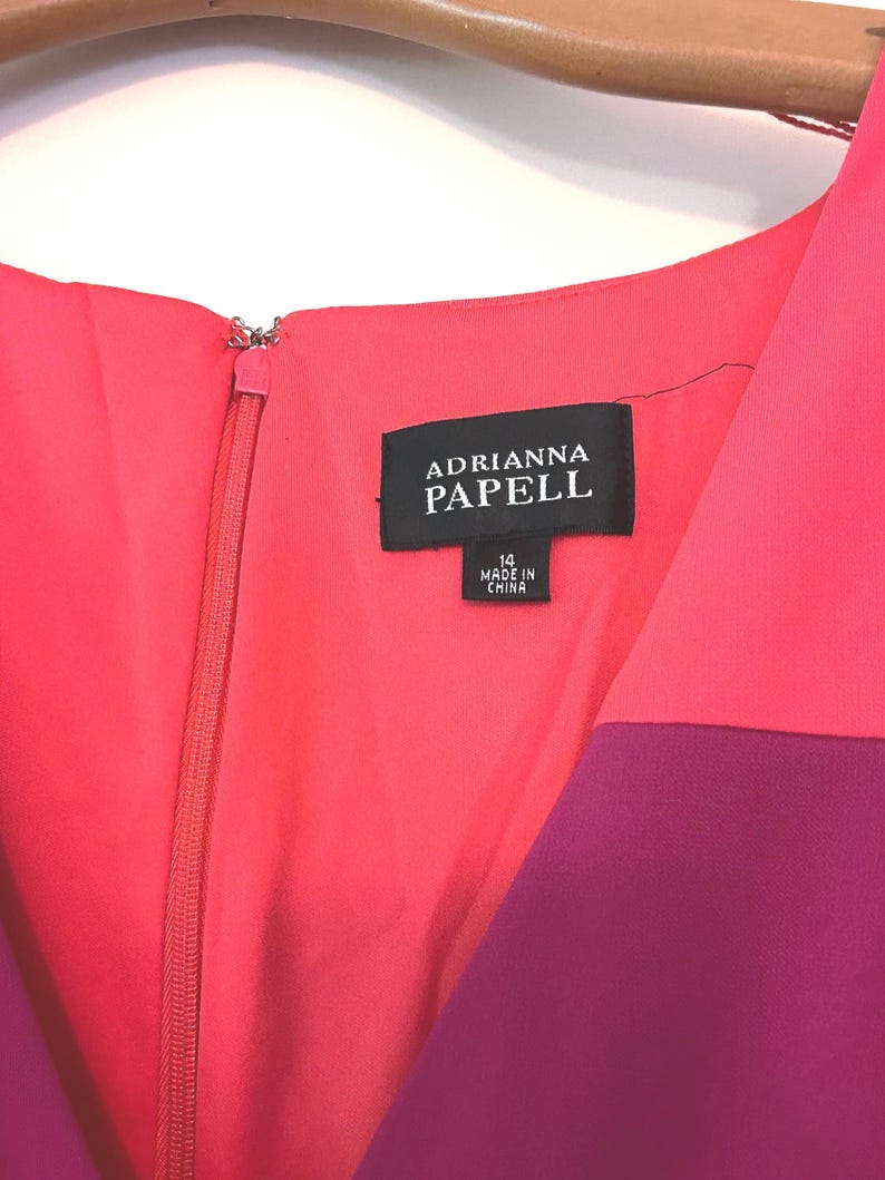 May include: A close-up of a color-blocked dress with a zipper. The dress features shades of pink and purple. The label reads "ADRIANNA PAPELL" and indicates a size 14, made in China.