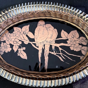 May include: An oval decorative tray featuring a gold-toned metal rim with a fluted edge and a woven wicker border. The center is a black glass inlay decorated with a light brown etched illustration of two birds perched on a flowering branch.