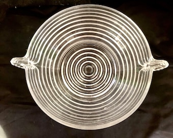 May include: A clear glass bowl with two handles. The bowl has a concentric circle design.
