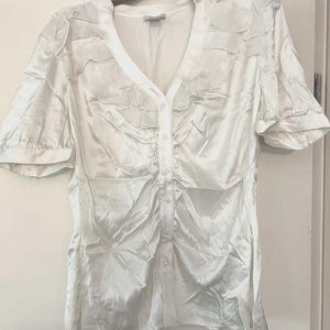 May include: A white short-sleeved blouse with a V-neckline and a button-down front. The garment features gathered fabric detailing along the front placket and shoulders, creating a textured, ruched appearance throughout the satin-like material.