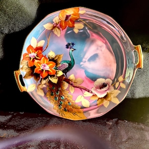 May include: A vintage porcelain serving dish with a peacock design. The dish is decorated with a peacock with a blue and green body, a gold tail, and orange flowers. The dish has a pink and blue background and gold trim.