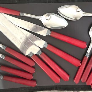 May include: A collection of silverware with red handles, including forks, knives, and spoons. The silverware is arranged on a dark surface, showcasing the contrast between the shiny metal and the vibrant red handles.